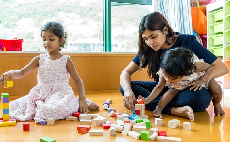 A teacher building with wooden blocks on the floor alongside two young children
